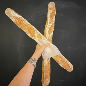 A person's hand holding two large baguettes in front of a dark chalkboard.