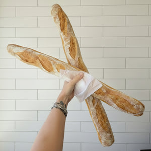 A person holds two large rustic baguettes crossed in an X shape in front of a white tiled wall