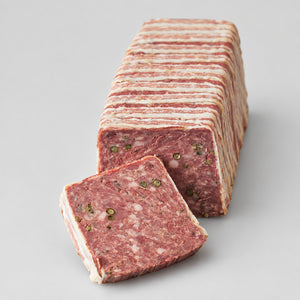 A sliced rectangular block of cured meat, likely salami or a similar marbled sausage, arranged on a dark wooden board with one slice placed in front. The meat shows pink-red tones, white fat bands, and small green herb flecks.