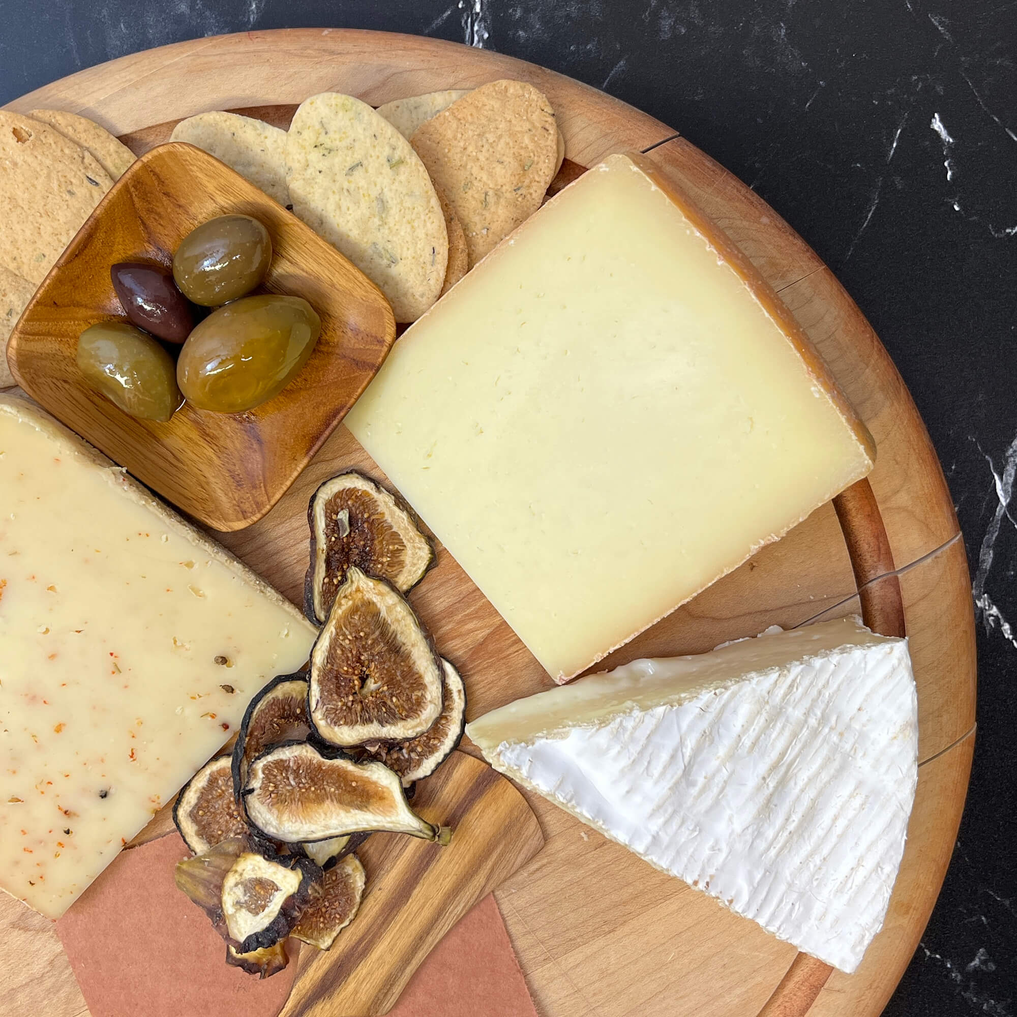 A spread of cheese, figs, olives, and crackers on a round wooden board.
